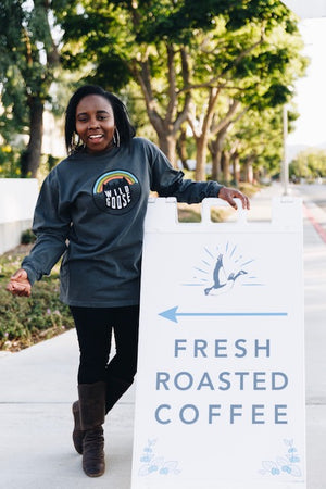 Girl posing in a Wild Goose Rainbow gray long sleeve, near our "Fresh Roasted Coffee" sign.